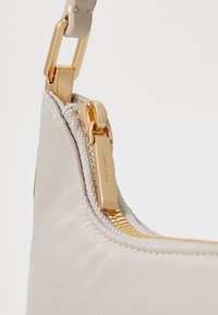 Geantă crossbody din piele bej moale, cu un fermoar auriu, accente texturate și un tag cu logo metalic, având un design elegant și o finisare netedă.