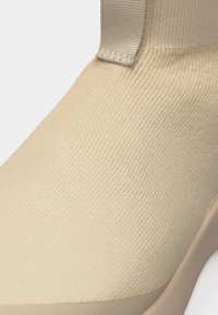 Κοντινό πλάνο ενός άνω μέρους παπουτσιού από μπεζ πλεκτό ύφασμα με θηλιά στο ύψος του αστραγάλου και λείο άκρο σόλας.