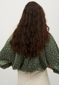 Blusa verde a pois con maniche larghe, realizzata in tessuto leggero, abbinata a pantaloni crema a vita alta. Capelli mossi e texturizzati visibili.