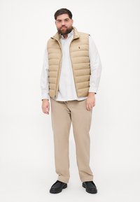 Béžový prešívaný vest s predným zipsom, s golierom, nosený cez bielu pruhovanú košeľu a béžové nohavice. Čierne hrubé topánky dokončujú outfit.