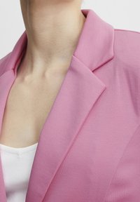 Pink blazer with a notched collar and smooth texture, featuring clean lines and a fitted design. Worn over a white ribbed top.