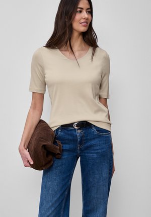 Mujer con camisa beige de manga corta y jeans azules sosteniendo una chaqueta marrón doblada, mirando hacia un lado contra un fondo liso.