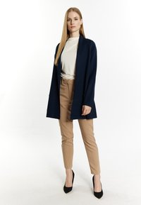 Cardigan lungo blu navy sopra una camicia bianca, abbinato a pantaloni beige e tacchi a punta neri; texture liscia e design dritto.