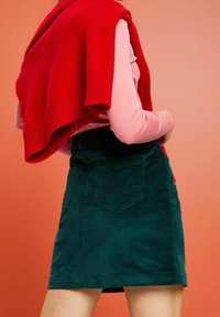 Groene corduroy rok met getextureerde strepen, voorzien van een aangesloten taille en twee achterzakken, gecombineerd met een rode gebreide trui over de schouders gedrapeerd.