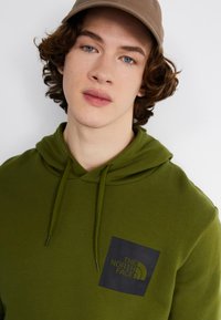 Felpa con cappuccio verde oliva, con cappuccio strutturato, tasca frontale e una patch quadrata scura con il logo "THE NORTH FACE" in lettere in grassetto.