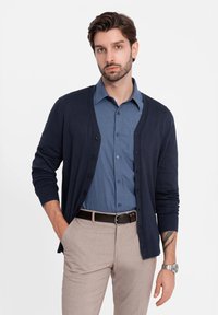 Cardigan navy scuro sopra una camicia azzurro chiaro con bottoni, abbinato a pantaloni beige. Presenta una texture liscia e una semplice rifinitura con bottoni.