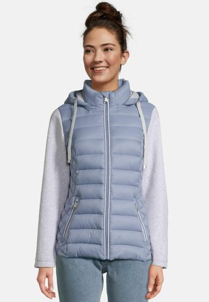 Young woman smiling wearing a light blue quilted sleeveless hooded vest over a gray long-sleeve shirt and jeans.