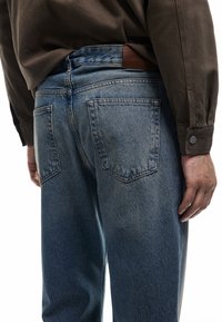 Vaqueros de mezclilla azul con un acabado ligeramente desgastado, que presentan un parche de cuero marrón en la cintura y bolsillos traseros tradicionales.