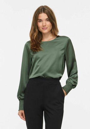 Femme aux longs cheveux bruns portant un chemisier en satin vert avec des poignets smockés et un pantalon noir taille haute, debout, une main dans la poche.