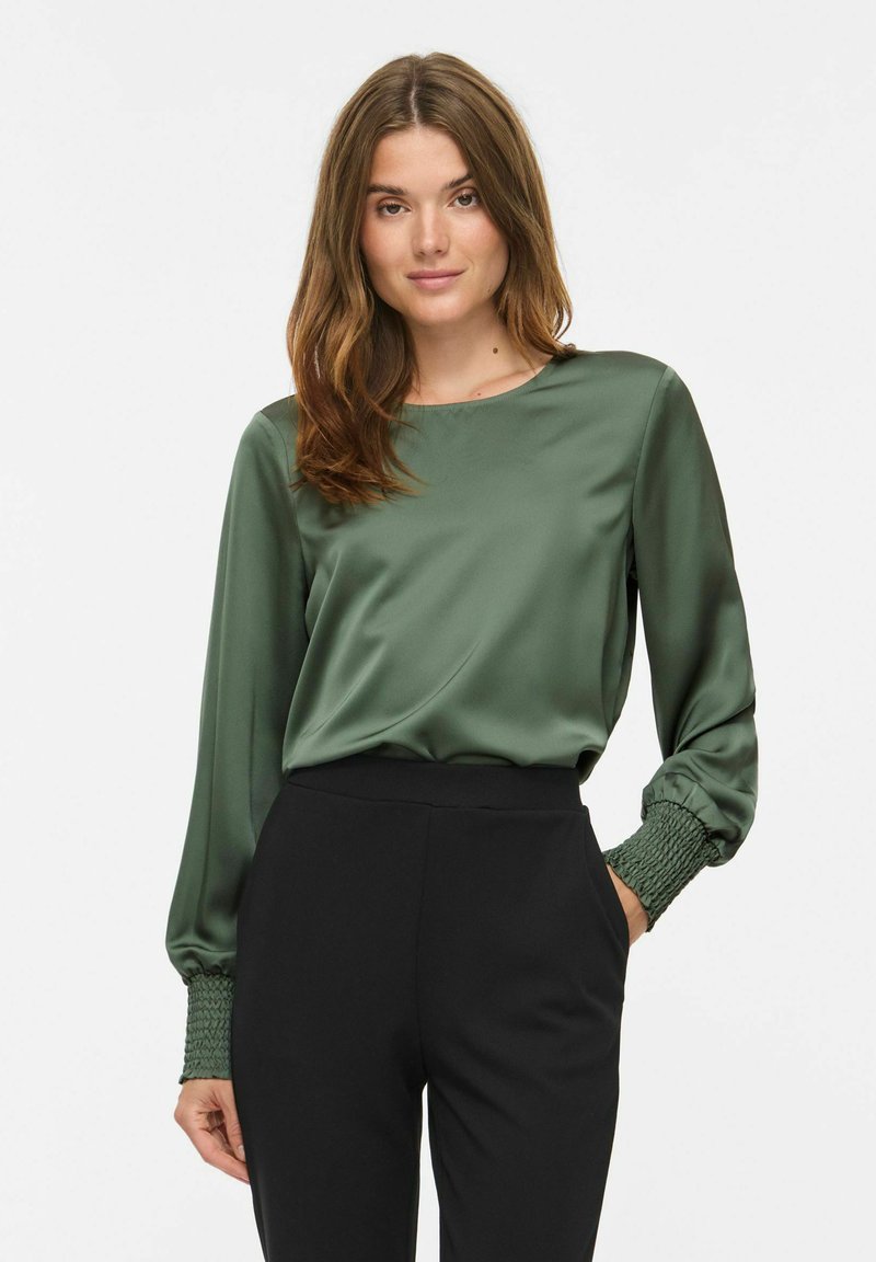 Femme aux longs cheveux bruns portant un chemisier en satin vert avec des poignets smockés et un pantalon noir taille haute, debout, une main dans la poche.