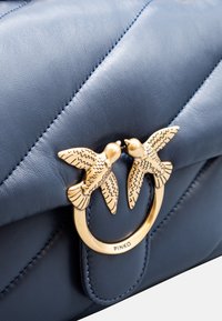 Borsa in pelle blu con texture trapuntata, dotata di una chiusura metallica dorata a forma di due uccelli che si affrontano. Logo visibile sulla chiusura.
