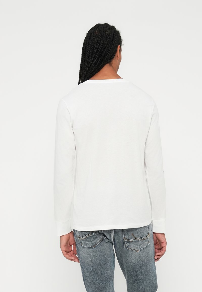 Camicia bianca a maniche lunghe in cotone, caratterizzata da un collo rotondo e una vestibilità rilassata. Dettagli di cucitura visibili sull'orlo e sui polsini.