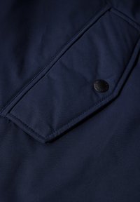 Tejido azul marino con una textura suave; cuenta con un gran bolsillo en ángulo y un detalle de botón a presión negro.