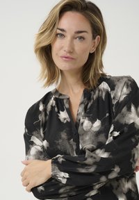 Blusa negra con estampado floral que presenta un escote con botones, mangas largas y puños elásticos. La tela tiene una textura suave y un patrón de acuarela.