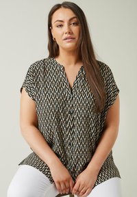Blusa de manga corta con patrón zigzag en negro y crema, con tapeta de botones y corte relajado, combinada con pantalones blancos.