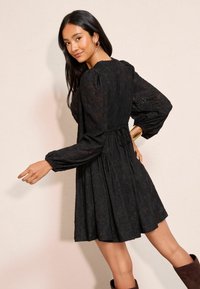 Musta mekko, jossa on pitkät puffihihat, kukkakuvioinen pitsirakenne, kokoontuva vyötärö ja valuva hame. Mallilla on päällään polviin ulottuvat saappaat ja asusteita.