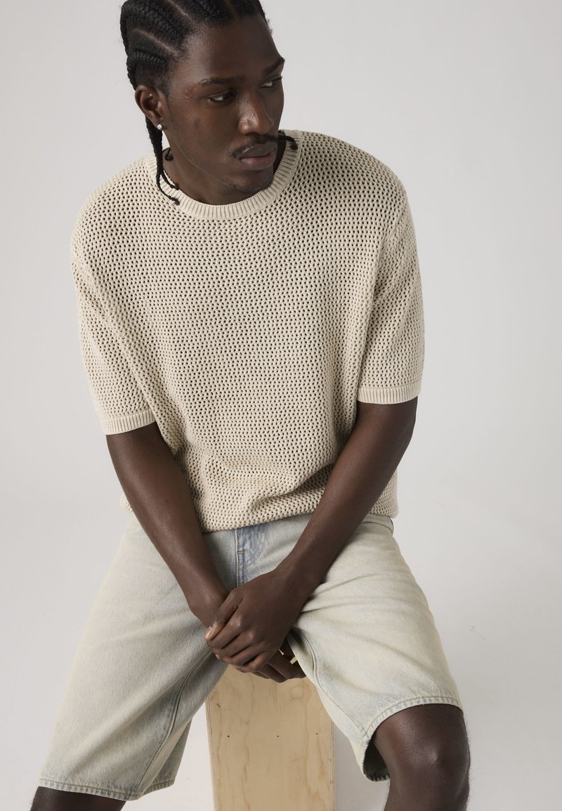 Homme aux cheveux tressés portant un t-shirt beige en maille à manches courtes et un short en jean clair, assis sur un bloc de bois, les mains jointes devant.