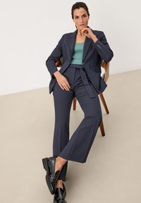  marineblazer en bijpassende broek van gladde stof, gecombineerd met een mintgroene geribbelde top. Zwarte schoenen met een glanzende afwerking.