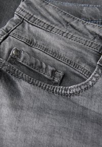 Grijze denim jeans hebben een achterzak met een metalen knoop, zichtbare stiksels en een zachte textuur. De stof heeft een lichte glans.