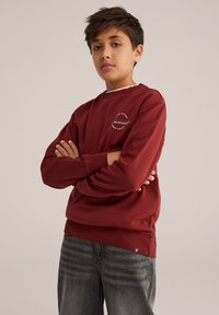 Röd bomulls sweatshirt med rund halsringning och ribbade muddar, med en logotyp tryckt på bröstet. Till det bärs grå denimjeans.