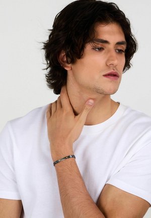 Braccialetto nero e oro con design intrecciato, caratterizzato da accenti piatti e finitura testurizzata, indossato al polso di una modella.