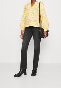 Personne portant un chemisier jaune à fleurs, un jean noir, des bottes à talons noires, et un sac à bandoulière noir sur un fond uni.