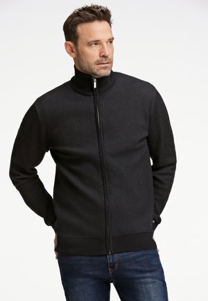 Chaqueta negra con cremallera y un cuello alto, fabricada con un tejido texturizado, puños y dobladillo acanalados. Presenta un sutil patrón de diamantes en toda su superficie.