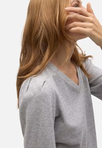 Top gris de manga larga con escote en V y pliegues sutiles en el hombro. Textura suave con un patrón jaspeado. Modelo parcialmente mostrado.