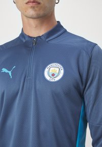Puma trening majica v temno modri barvi z visokim ovratnikom, pol-zipom, logotipom Manchester City in subtilnim teksturiranim vzorcem. Aqua detajli na rokavih.