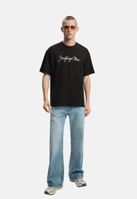 T-shirt nera oversize con testo in bianco, abbinata a jeans svasati azzurri chiari e sneakers grigie. Il modello indossa occhiali da sole rotondi.