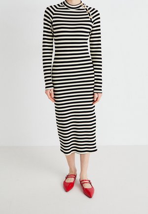 Žena nosící černobílé midi šaty s dlouhým rukávem a horizontálními pruhy, s knoflíky na ramenou a červenými špičatými botami Mary Jane.