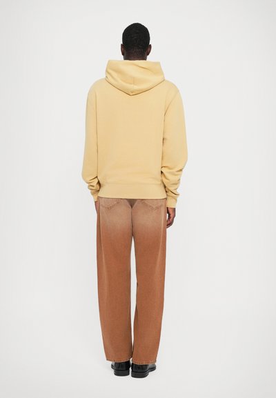 Mann som står med ryggen til, iført en beige hettegenser, løse brune falmede jeans og svarte sko mot en enkel hvit bakgrunn.