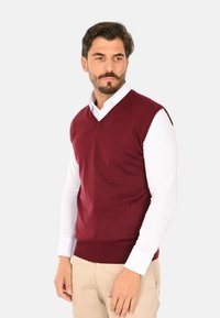 Canotta bordeaux senza maniche realizzata in tessuto a maglia, con un design a scollatura a V, indossata sopra una camicia bianca a maniche lunghe.