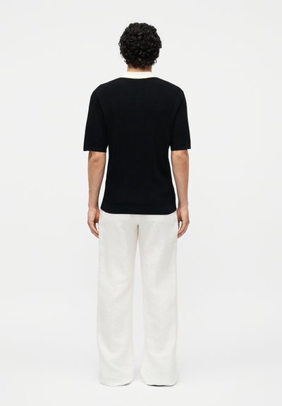 Homme aux cheveux bouclés et foncés, debout de dos, portant une chemise noire à manches courtes et un pantalon blanc à jambes larges sur un fond uni.