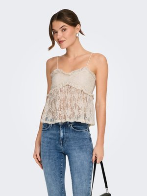 Mujer con camisola de encaje beige con tirantes finos y vaqueros azules, sosteniendo un bolso negro a su lado.