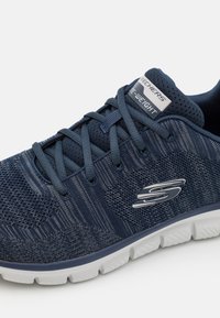 Zapatilla deportiva azul marino que cuenta con una parte superior de malla, patrones texturizados, cierre de cordones y una suela de goma blanca con el logotipo de Skechers en el costado.