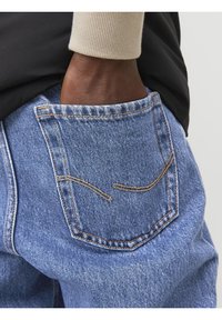 Jeans in denim con vita alta e lavaggio blu sbiadito. Presentano tasche posteriori con cuciture a contrasto e una texture morbida, mostrati con una mano nella tasca.