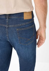 Jeans in denim blu scuro con una toppa in pelle beige sulla vita e cuciture distintive sulla tasca posteriore. Texture liscia.