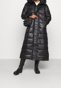 Abrigo largo acolchado negro con capucha, presenta cierre de cremallera, diseño acolchado y una textura brillante. Combinado con botas negras hasta la rodilla y un bolso pequeño.