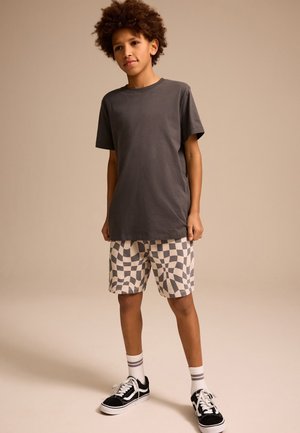 Dreng med krøllet hår, stående iført en mørkegrå t-shirt, ternede shorts, hvide sokker og sorte sneakers foran en ensfarvet baggrund.