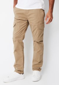 Khaki cargobukser lavet af bomuldsblandingsstof; har flere sidelommer; tilspidsede ben og elastiske manchetter. Bæres med hvide sneakers.