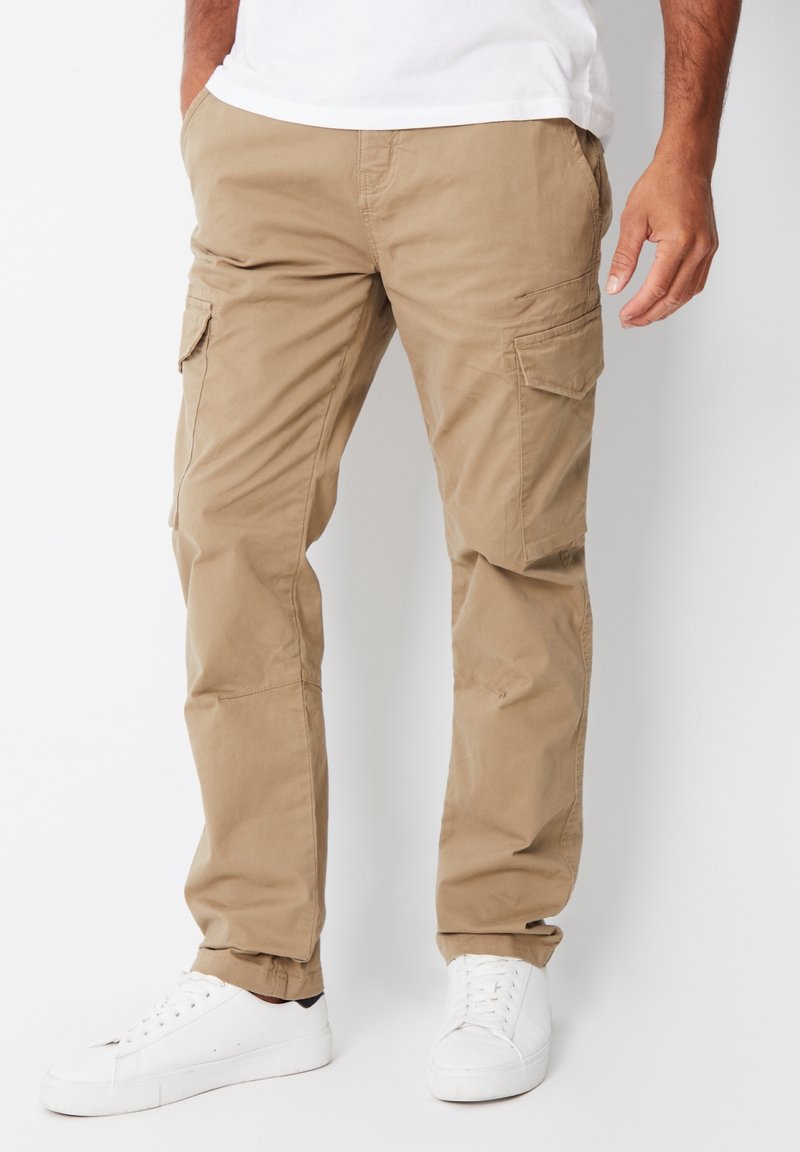 Khaki cargobukser lavet af bomuldsblandingsstof; har flere sidelommer; tilspidsede ben og elastiske manchetter. Bæres med hvide sneakers.