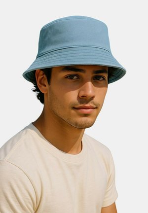 Młody mężczyzna z lekkim zarostem, noszący jasnoniebieski kapelusz bucket hat i gładką kremową koszulkę, patrzący prosto przed siebie o neutralnym wyrazie twarzy.