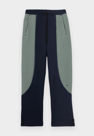 Nike trenirke s dvobarvnim dizajnom v zeleni in mornarsko modri barvi, s stranskimi žepi, vrvico za prilagajanje pasu in nastavljivimi robovi.