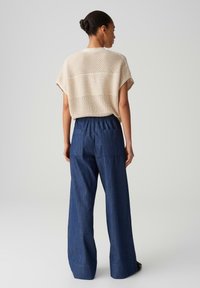 Beige open-knit sweater with drop shoulders over dark blue wide-leg denim trousers featuring a relaxed waistband and two back pockets.