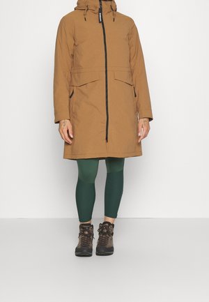 Personne portant un parka à capuche marron avec des poches avant, des leggings verts et des chaussures de randonnée marron, debout devant un fond blanc uni.