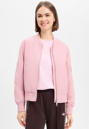 Jonge vrouw glimlachend, draagt een lichtroze bomberjack met rits, een bleekroze shirt en donkerbruine Adidas trainingsbroek met witte strepen.