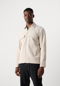 Lichtbeige corduroy jack met een voorste rits, twee borstzakken en lange mouwen. Getextureerde stof, casual ontwerp, gedragen met donkere jeans.