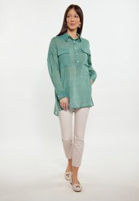 Camicia di lino teal con design a bottoni, maniche lunghe e due tasche sul petto. Abbinata a pantaloni fitted beige chiaro e scarpe basse beige.