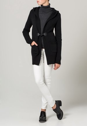 Cardigan nero lavorato a maglia con cappuccio, dotato di chiusura a nastro e due tasche frontali, abbinato a pantaloni bianchi aderenti e scarpe nere.